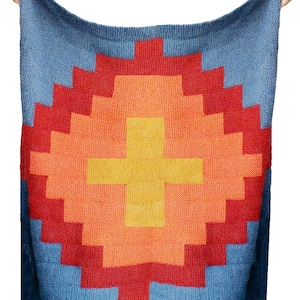 May include: A knitted blanket with a geometric design. The blanket features a central cross shape in yellow, surrounded by layers of orange and red, set against a blue background. The blanket is made of a soft, textured material.