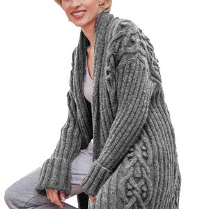 May include: A grey, long-sleeved cardigan with a shawl collar. The sweater features a cable knit design on the sleeves and front panels. The cuffs are ribbed. The model is wearing a grey undershirt and grey tracksuit bottoms.