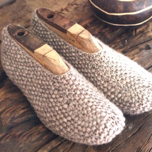 May include: A pair of beige knitted slippers with wooden shoe trees inside. The slippers are on a wooden surface.