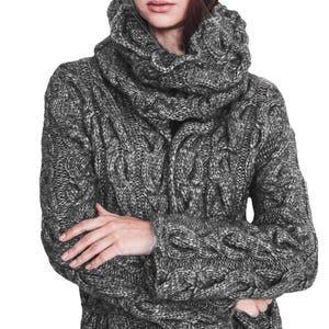 Puede incluir: Un suéter gris de punto trenzado con cuello alto. El suéter tiene una apariencia gruesa y texturizada con intrincados patrones tejidos. Las mangas son largas y el diseño sugiere una prenda cálida y acogedora.