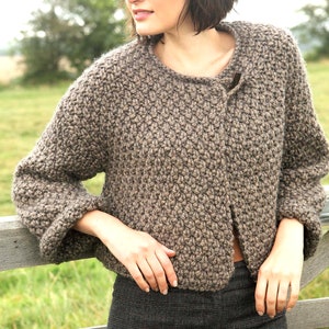 以下が含まれることがあります： ボタン留めの茶色のニットカーディガン。カーディガンは、質感のあるニット模様で、ショートカットの茶髪の女性が着用しています。