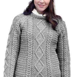 Puede incluir: Un suéter gris de manga larga con un complejo patrón de tejido de cable con trenzas verticales y formas de diamante. El tejido tiene una apariencia texturizada con sutiles motas plateadas en todo el material, cuello redondo y puños acanalados.