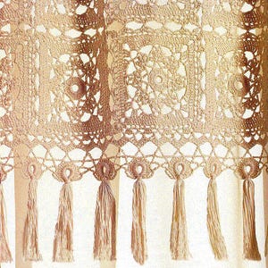 Op de afbeelding: Een close-up van een beige gehaakt gordijn met een gedetailleerd openwerkontwerp. Het gordijn heeft vierkante en cirkelvormige patronen, met een geschulpte rand en lange kwasten die aan de onderkant hangen. De textuur is ingewikkeld en delicaat.