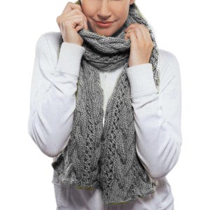 Puede incluir: Una bufanda de punto gris con un patrón de trenza. La bufanda la lleva puesta una persona con un suéter blanco.