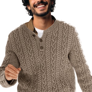 May include: A tan cable-knit sweater with a button-up neckline. The sweater has a textured pattern and long sleeves. The person is wearing a white shirt underneath the sweater.
