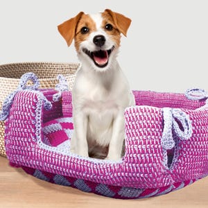 May include: A happy Jack Russell Terrier sits in a handmade, crochet dog bed. The bed is a vibrant pink and white, with blue accents and decorative ties. A woven basket is in the background.