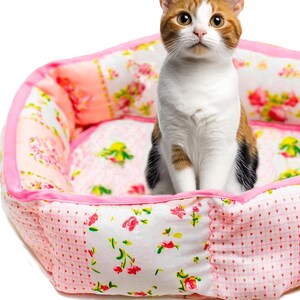 Puede incluir: Un gato calic&oacute; est&aacute; sentado en una cama para mascotas rosa y con estampado floral. La cama tiene un ribete de sat&eacute;n rosa y est&aacute; hecha de tela acolchada. El gato tiene pelaje naranja, blanco y negro y mira directamente a la c&aacute;mara.