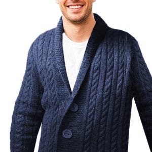 Op de afbeelding: Een donkerblauw vest met kabelgebreid patroon, voorzien van een sjaalkraag en twee grote bijpassende knopen aan de voorzijde. Het kledingstuk wordt gedragen over een effen wit T-shirt en toont een gestructureerd breipatroon met verticale kabeldetails.