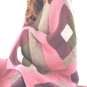 Könnte beinhalten: Eine gestrickte Decke mit einem geometrischen Muster aus rechteckigen Blöcken in den Farben Pink, Dunkellila, Olivgrün und Creme. Das Textil weist eine durchgehende kraus rechts gestrickte Textur auf, die ein weiches und strukturiertes Erscheinungsbild erzeugt.