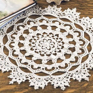 May include: A white crocheted doily with a delicate, intricate design. The doily is circular and has a center with a smaller, more tightly woven pattern. The doily is laid out on a wooden surface.