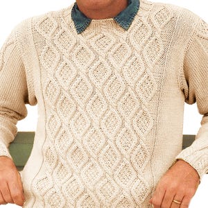 May include: Cream-colored knit sweater with a diamond pattern. The sweater has a crew neck and long sleeves with cable knit detailing. The person is wearing a denim collared shirt underneath the sweater.