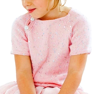 May include: A short-sleeved light pink knit sweater featuring scattered silver sequins throughout the fabric. The garment has a round neckline and two white buttons positioned on the right shoulder area, providing a subtle decorative detail.