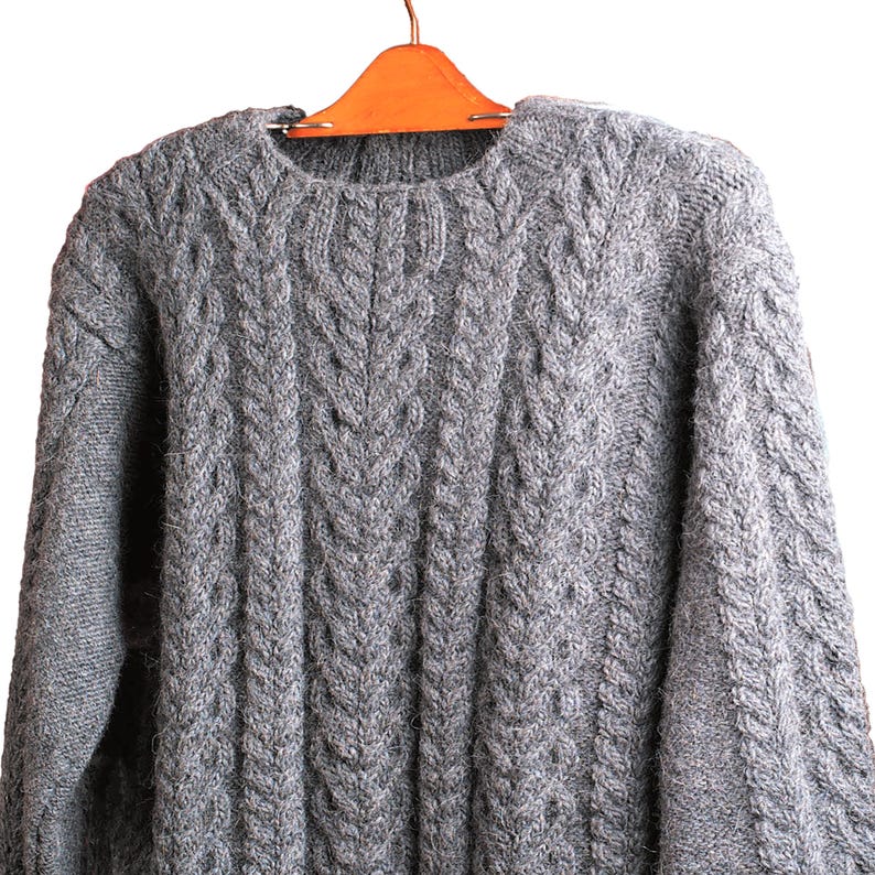 May include: A gray cable knit sweater hanging on a wooden hanger. The sweater has a classic crew neck and a textured pattern.