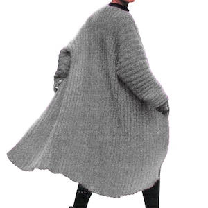 Op de afbeelding: Een lange, grijze, geribbelde gebreide cardiganjas. De jas heeft een vloeiend, oversized ontwerp en wordt gedragen met zwarte laarzen en een bijpassende grijze beanie. De persoon draagt een zonnebril en kijkt over zijn schouder.