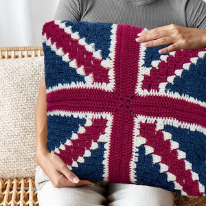 Pode incluir: Uma almofada quadrada de crochê com o design da bandeira britânica. A almofada tem um fundo azul marinho com uma cruz bordô e detalhes brancos. A almofada está sendo segurada por uma pessoa.