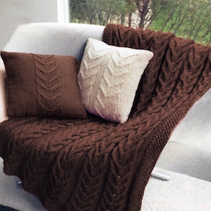 May include: A brown cable knit throw blanket and two pillows, one brown and one white, are arranged on a white couch. The throw blanket and pillows have a cable knit pattern.