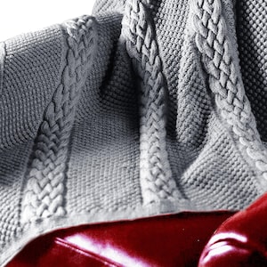 Puede incluir: Una manta de punto de cable gris con un patr&oacute;n texturizado. La manta est&aacute; drapeada sobre un sof&aacute; de cuero rojo.