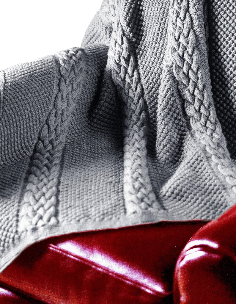 Puede incluir: Una manta de punto de cable gris con un patr&oacute;n texturizado. La manta est&aacute; drapeada sobre un sof&aacute; de cuero rojo.