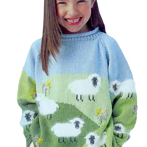 May include: A light blue knitted sweater featuring a pastoral scene with white sheep grazing on a green hillside under a blue sky. The sweater has long sleeves and is decorated with yellow and purple flowers.