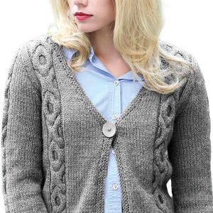 May include: A gray, cable-knit cardigan with a V-neck and a large, round button closure. The sweater features intricate cable patterns along the front and sleeves. The model is wearing a light blue collared shirt underneath.