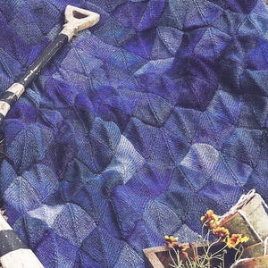 Op de afbeelding: Een gebreide deken in blauw- en paars tinten met een gestructureerd, geometrisch patroon. Een kleine schop en een houten plantenbak met gele bloemen staan op de voorgrond. De deken is opgevouwen en lijkt buiten te zijn.