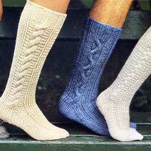 May include: Four pairs of knee-high knitted socks in various colors: gray, cream, blue, and off-white. Each sock features a cable knit design. The socks are displayed on a green surface, showcasing the different textures and patterns.