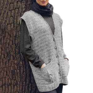 Peut inclure: Un gilet sans manches en tricot gris clair avec une encolure en V, une fermeture boutonnée à l'avant avec six boutons et deux grandes poches plaquées. Le gilet est porté sur un haut à manches longues et une écharpe gris foncé texturés.