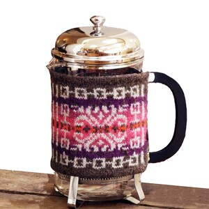 Könnte beinhalten: Eine Glas-French-Press-Kaffeemaschine mit einer gestrickten Hülle. Die Hülle ist braun mit einem geometrischen Muster in Pink, Lila und Weiß.