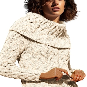 Pode incluir: Camisola de malha creme com gola larga e dobrada. A camisola apresenta um padrão texturizado e mangas compridas. A pessoa está segurando óculos de sol. A camisola é uma peça de roupa aconchegante e quente.