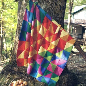 Peut inclure: Une couverture tricotée colorée avec un motif géométrique de triangles dans des tons de rouge, orange, jaune, vert, bleu et rose. La couverture est drapé sur un tronc d'arbre dans un cadre boisé.