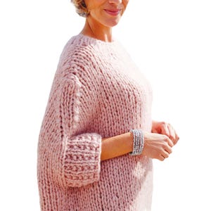 May include: A light pink, chunky knit sweater with a crew neck and three-quarter sleeves. The sleeves have a ribbed cuff. A silver bracelet is visible on the wrist. The sweater is made of a thick, soft yarn.