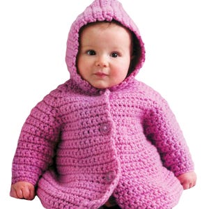 May include: A baby wearing a handmade, hooded, pink crochet jacket. The jacket has a button closure and a textured stitch pattern. The baby is looking directly at the camera.