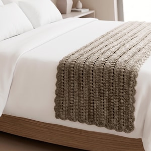May include: A taupe-colored crochet bed runner featuring a textured pattern of vertical bobble stitches and a scalloped edge. The runner is draped across a white bedspread, showcasing a detailed knit construction with a consistent earthy tone.