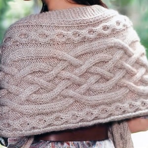 Può includere: Uno scialle di maglia beige con un motivo a treccia. Lo scialle ha un design semplice con un motivo ripetitivo di trecce intrecciate.