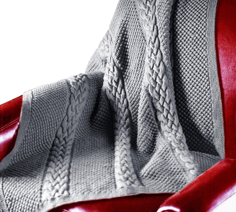 Puede incluir: Una manta de punto de cable gris con un patr&oacute;n texturizado. La manta est&aacute; drapeada sobre un sill&oacute;n rojo.