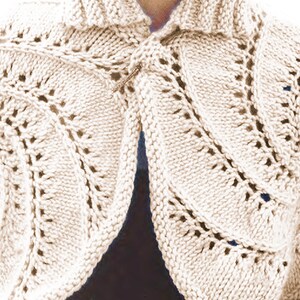 May include: A cream-coloured knitted shrug with a leaf pattern. The open-front design features a collar and is fastened with a decorative pin. The knitwear has a textured appearance with openwork details, suitable for layering.