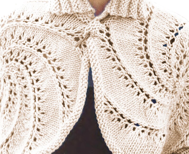 May include: A cream-coloured knitted shrug with a leaf pattern. The open-front design features a collar and is fastened with a decorative pin. The knitwear has a textured appearance with openwork details, suitable for layering.