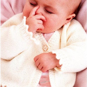 May include: A sleeping baby wearing a cream-colored, knitted cardigan with scalloped edges and a single button closure. The baby is nestled in a pink blanket and wearing a pink onesie.