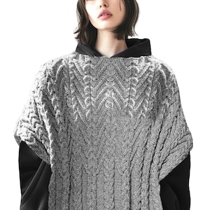 Peut inclure: Un poncho en tricot de câble gris avec un motif géométrique. Le poncho est porté sur un haut noir. Le design tricoté présente un aspect texturé et tissé. Le modèle porte un haut à capuche noir sous le poncho.
