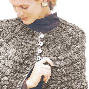 Op de afbeelding: Een bruine gebreide cape met een kabelbreipatroon en witte knopen. De cape heeft een ronde halslijn en een losse pasvorm.