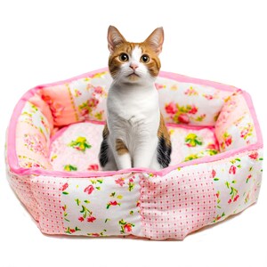 Puede incluir: Un gato calic&oacute; est&aacute; sentado en una cama para mascotas rosa y blanca con estampado floral. La cama tiene un ribete rosa y un dise&ntilde;o de patchwork con estampados florales y de lunares. El gato tiene pelaje naranja, blanco y negro.