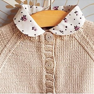 May include: A beige knitted cardigan with a white collared shirt underneath. The shirt has a small floral pattern in purple and black.