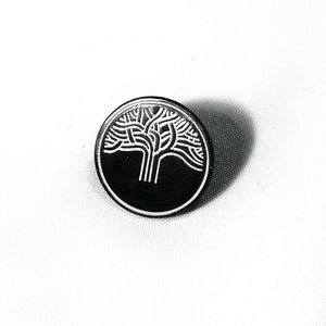 May include: A silver-coloured pin featuring a black and white tree design. The tree is stylised and has a thick trunk with many branches.