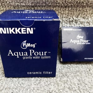 May include: Two dark blue rectangular boxes for a Nikken PiMag Aqua Pour gravity water system. The larger box is labeled ceramic filter, and the smaller box is labeled microsponge. Both boxes feature white text and the brand logo on the front.