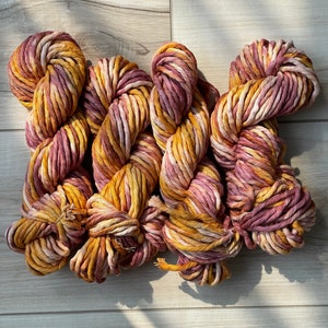 May include: Four skeins of chunky yarn in shades of yellow, pink, and cream. The yarn is tightly wound and has a textured appearance. The skeins are arranged on a light wood surface, with shadows adding depth.