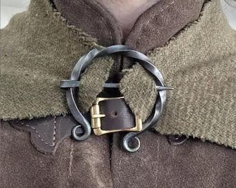 May include: A dark gray twisted metal circular brooch with a pointed pin, securing a brown leather strap with a brass buckle. The brooch fastens a textured olive green fabric layer over a dark brown suede garment.