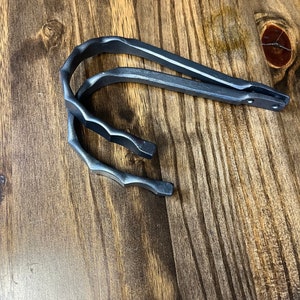 May include: Two dark gray metal stirrups with a curved design. The stirrups have a textured, hand-forged appearance and are set against a wooden background. The stirrups are likely for equestrian use.