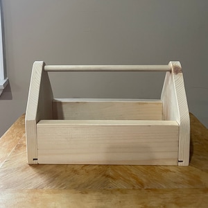 May include: A plain wooden toolbox with a handle. The toolbox is made of unfinished wood and has a rectangular shape.