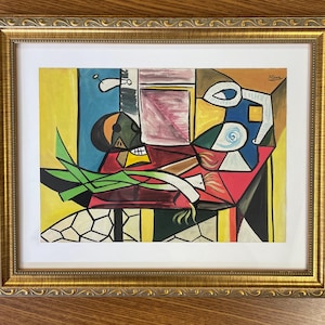 May include: A framed cubist-style painting featuring a vibrant composition of geometric shapes, including a red table surface, green leeks, and abstract forms in yellow, blue, and black. The artwork is signed Picasso and encased in an ornate gold frame.