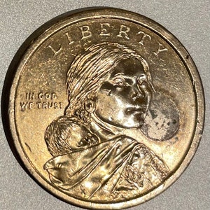 Può includere: Una moneta da un dollaro americano dorata con le parole "Liberty" e "In God We Trust" incise su di essa. La moneta presenta il profilo di una donna con un bambino sulla schiena.